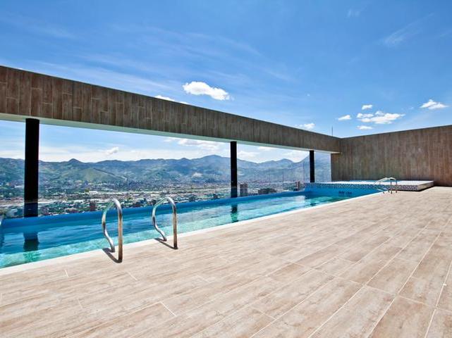 An infinity rooftop pool is situated on a sunlit terrace, offering expansive views of the surrounding city valley and mountains. Smooth tiles surround the pool, and modern lounge chairs are positioned nearby, creating a relaxing atmosphere for guests to enjoy the scenery.