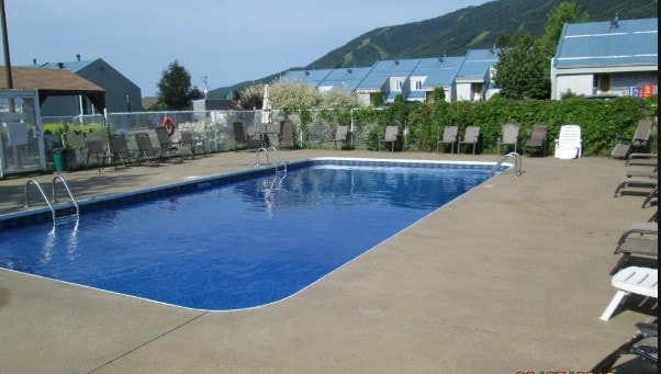 An outdoor heated pool is surrounded by a spacious deck, featuring multiple lounge chairs arranged neatly around it. Lush greenery and distant hills are visible in the background, providing a serene setting for relaxation.