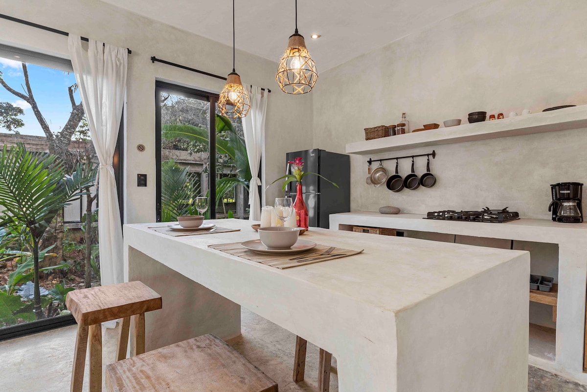 The modern kitchen features a large, minimalistic table with two dining sets positioned on rustic stools. Natural light fills the space through expansive glass doors, while lush greenery is visible outside. Various kitchen utensils and appliances are neatly arranged on open shelves.