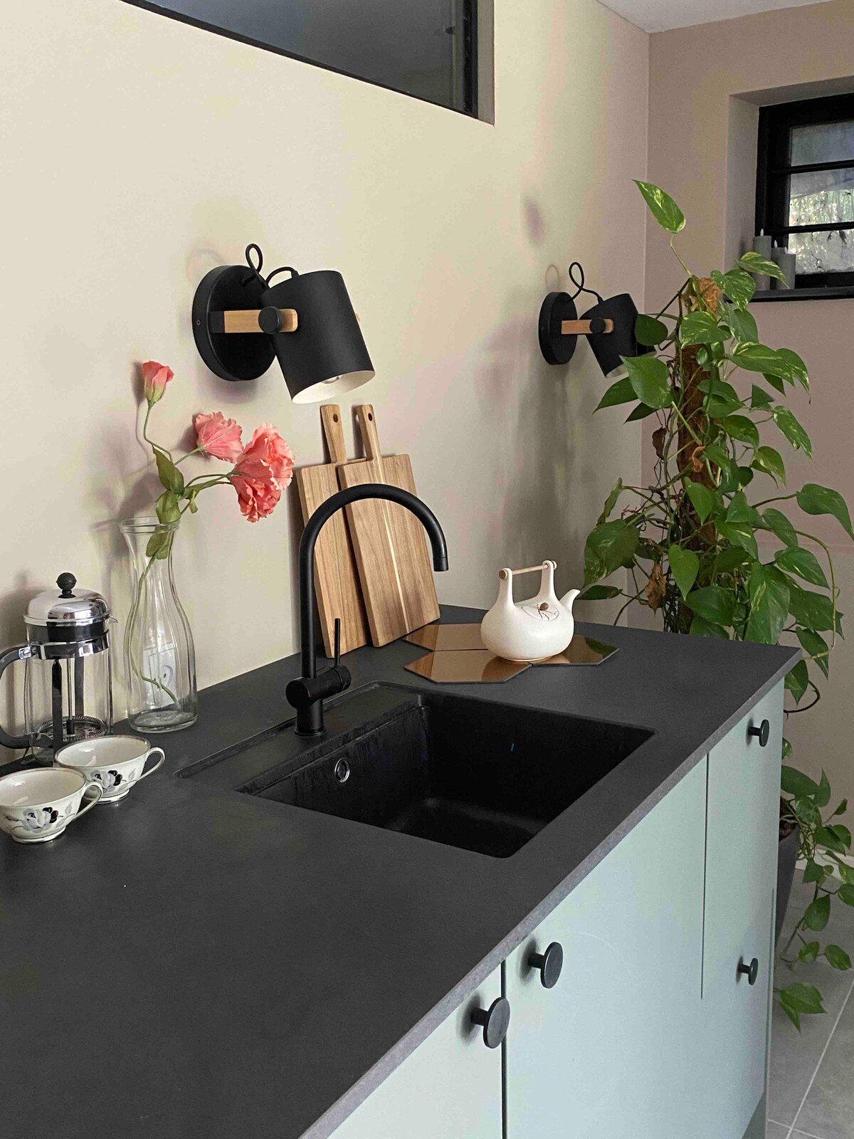 The kitchen area features a sleek black countertop paired with light green cabinetry. A modern faucet is situated above a small sink, while two wall sconces provide soft illumination. Kitchenware, including a French press, is arranged nearby, enhanced by fresh flowers and a decorative plant.