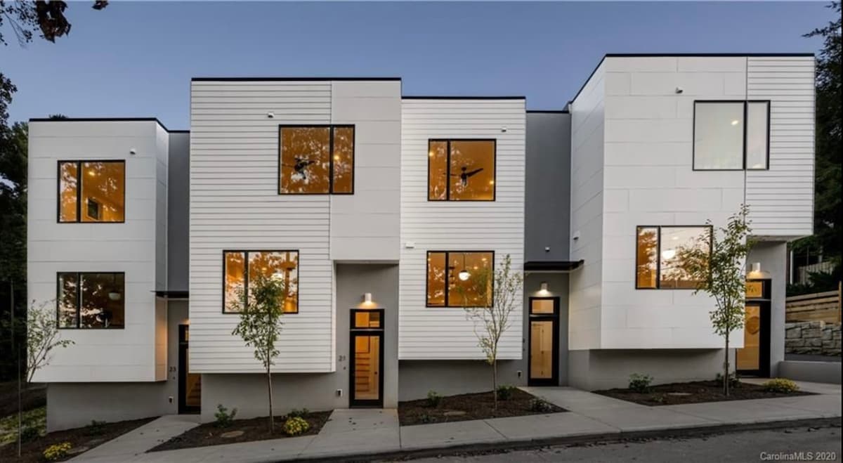 A modern multi-unit condo building features a clean white facade with large windows reflecting warm evening light. Landscaping includes small trees and a well-maintained path leading to the entrances, which are framed by simplified architectural details.