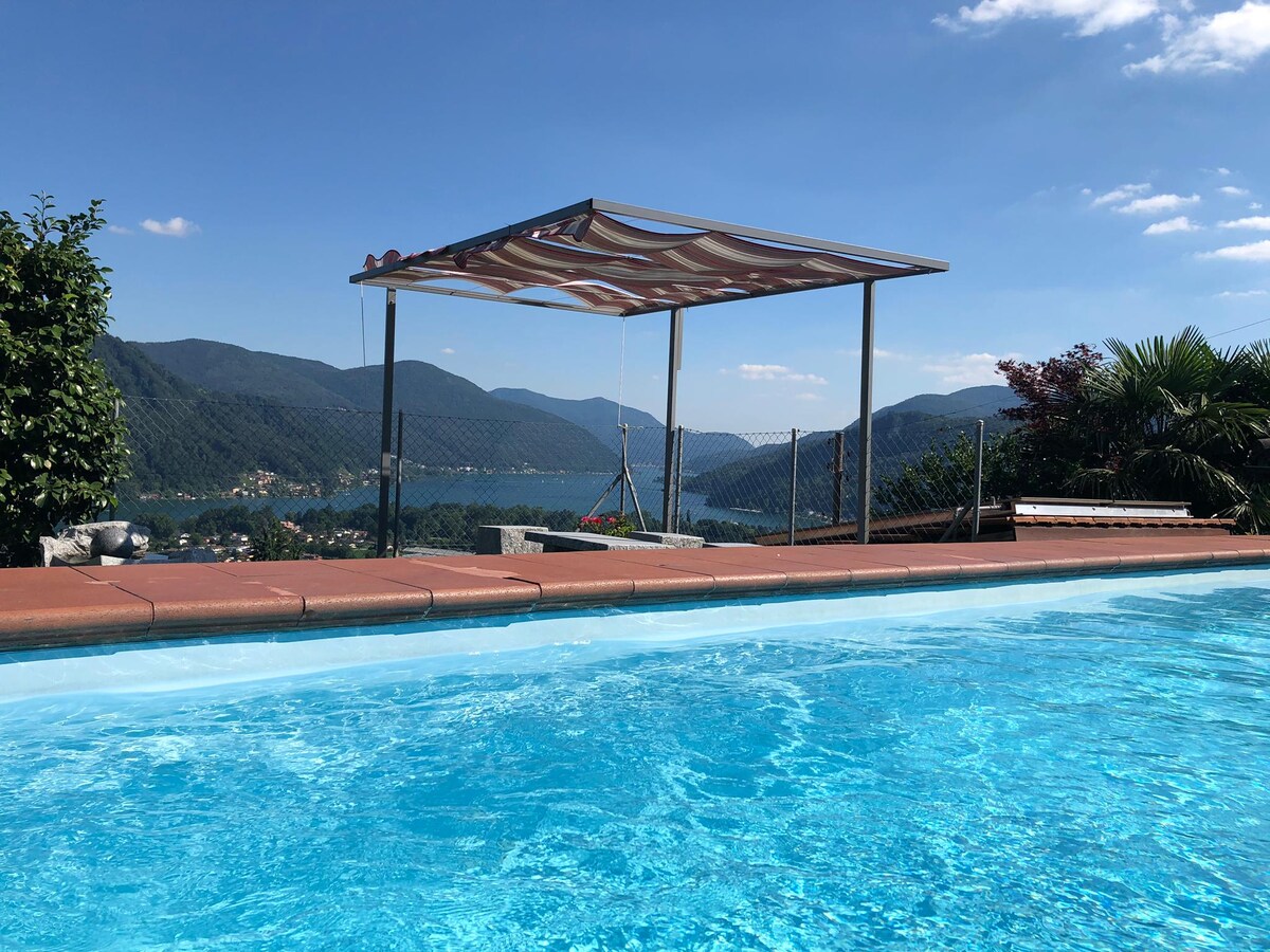 An outdoor swimming pool reflects the clear blue sky, framed by a sunshade structure. Lush greenery is visible at the pool's edge, while distant mountains and a tranquil lake provide a serene backdrop.