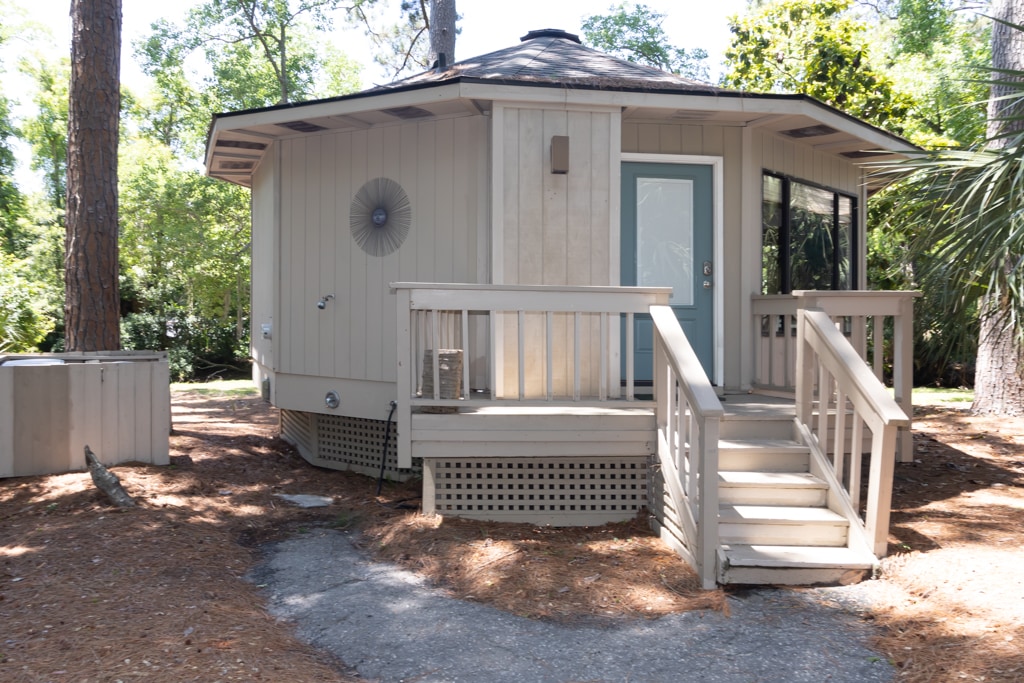 A charming cottage exterior is shown with a wrapped entryway and steps leading to the front door. Surrounding greenery and trees create a serene environment. A fan is mounted on the wall, and the large window allows for natural light into the living space.