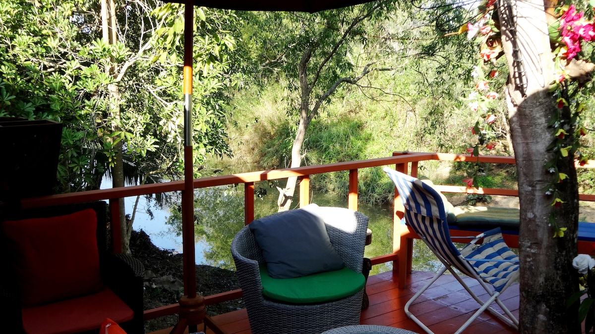 A covered outdoor seating area is surrounded by lush greenery, overlooking a serene river. Comfortable chairs and a striped lounge chair are arranged on a wooden deck, inviting relaxation and offering a peaceful view of the natural surroundings.