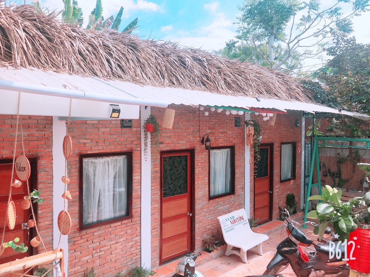 A row of three private rooms is presented, featuring rustic brick walls and wooden doors. Each room has white curtains allowing natural light to enter. The roof is adorned with thatch, and potted plants add greenery, creating a welcoming outdoor space.