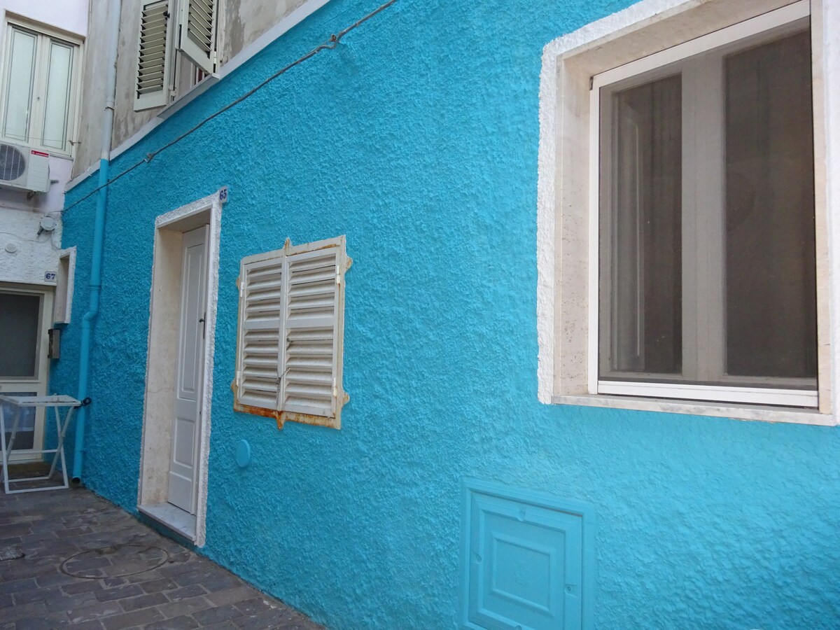 A vibrant blue exterior wall of the building is visible, featuring a simple doorway and a window. The textures of the wall stand out, complemented by a wooden shutter and a decorative panel. Light-colored accents create a neat appearance against the vivid background.