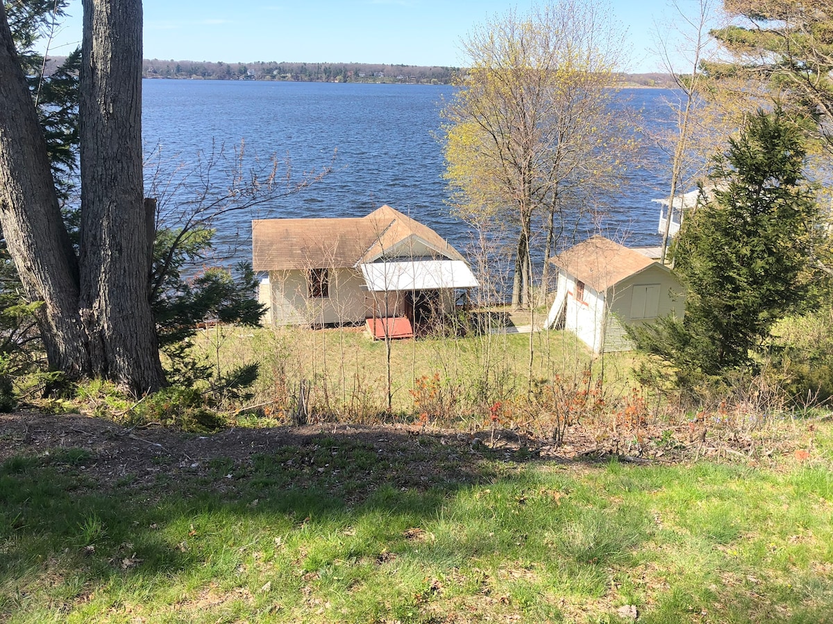 A serene view of a rustic cottage and boathouse is framed by lush greenery. The structures are nestled on a grassy lot, with Lake Michigan visible in the background. Trees provide natural shade, enhancing the peaceful lakeside setting.