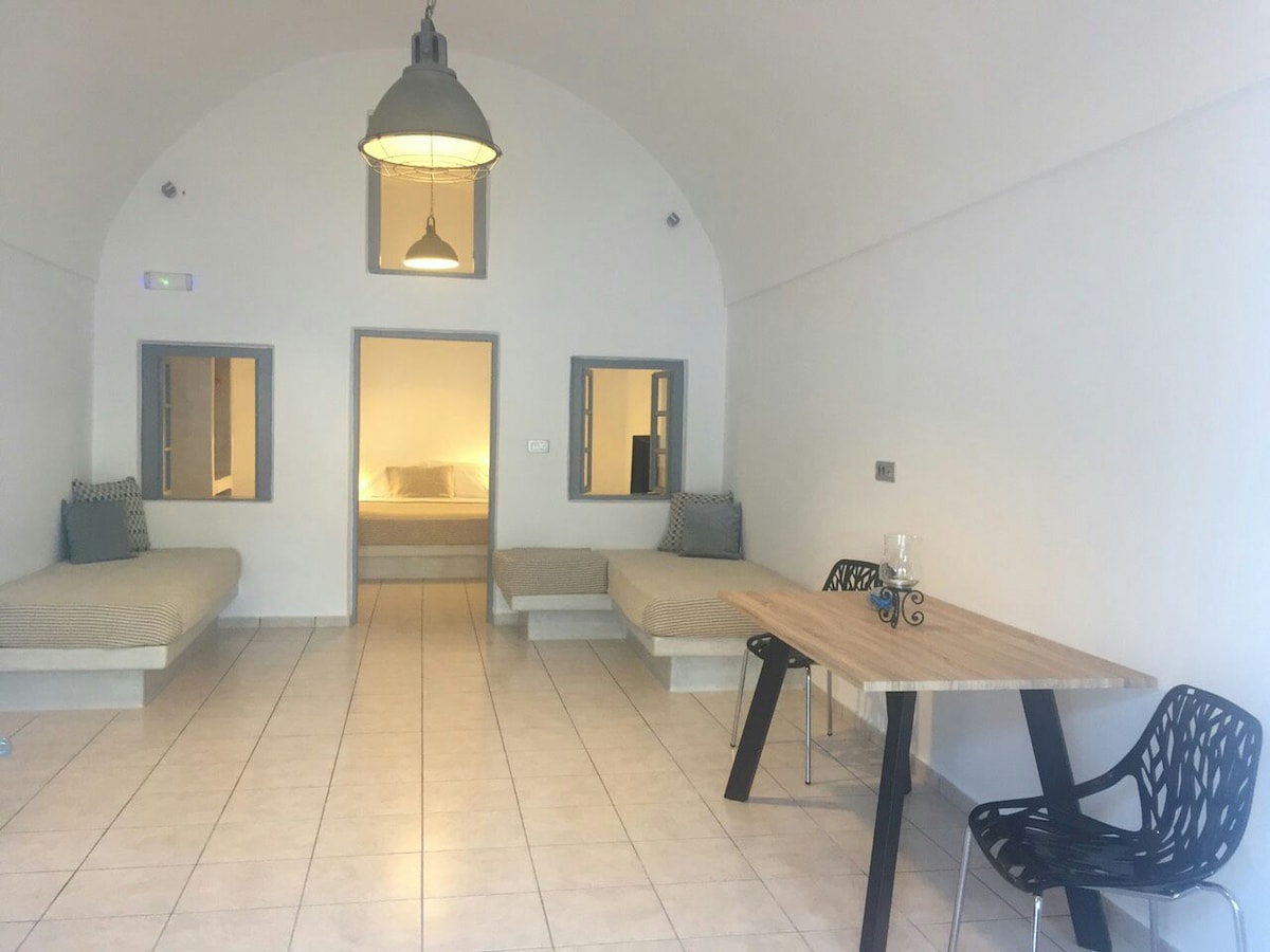 The spacious sitting room features two built-in seating areas with neutral-colored cushions and a wooden dining table surrounded by black chairs. Large windows provide natural light, and an overhead pendant light enhances the airy interior of the traditional cave house.