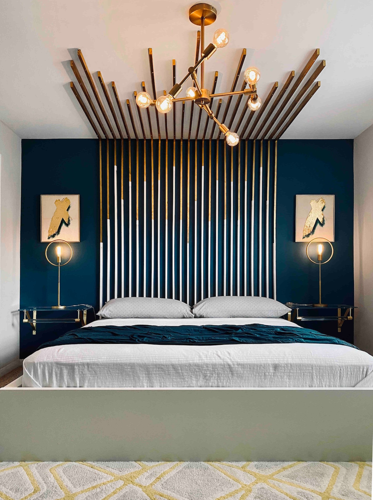 A modern bedroom features a stylish metal headboard, framed by vertical lines in a deep blue hue. A spacious bed is dressed in a white duvet and dark teal accents. Minimalist side tables with round lamps provide soft illumination, enhancing the room's contemporary design.