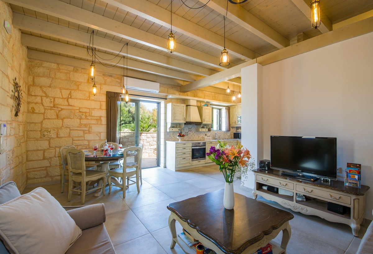 An open living area features stylish wood and stone accents, illuminated by pendant lighting. A comfortable sofa is positioned near a coffee table with a flower vase. A dining table for four is visible, along with a modern kitchen and large windows offering views of the outdoors.