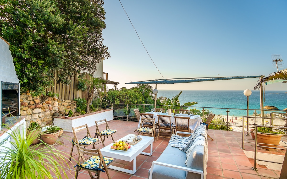 A spacious outdoor patio is highlighted by comfortable seating, including wooden chairs and a large table. A wood-fired BBQ is visible, surrounded by lush greenery. The ocean view captures the serene beach, with clear skies creating a relaxed ambiance.