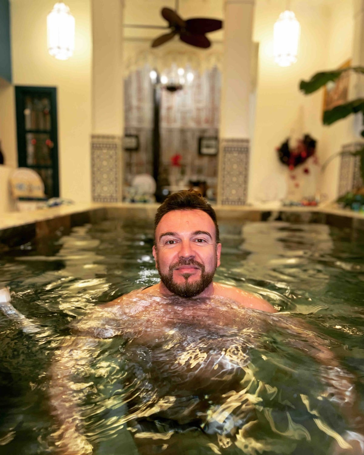 An indoor swimming pool is visible, featuring clear, warm water reflecting soft lighting. Surrounding the pool, elegant tiles and potted greenery create a serene atmosphere. Light filters through sheer curtains, illuminating the space while maintaining privacy.