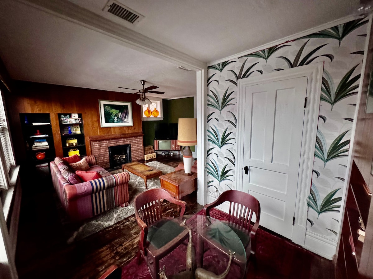 A living area features a striped sofa, wooden coffee tables, and a fireplace adorned with a large piece of art. A glass dining table with two chairs is positioned nearby. Vibrant botanical wallpaper decorates one wall, while natural light enters through the windows.