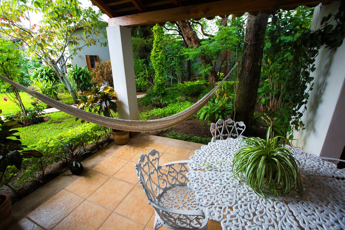 A private terrace is seen with a light-colored table surrounded by intricately designed chairs. A hammock hangs nearby, offering a relaxed space, while lush greenery is visible beyond, creating a tranquil environment in the garden.