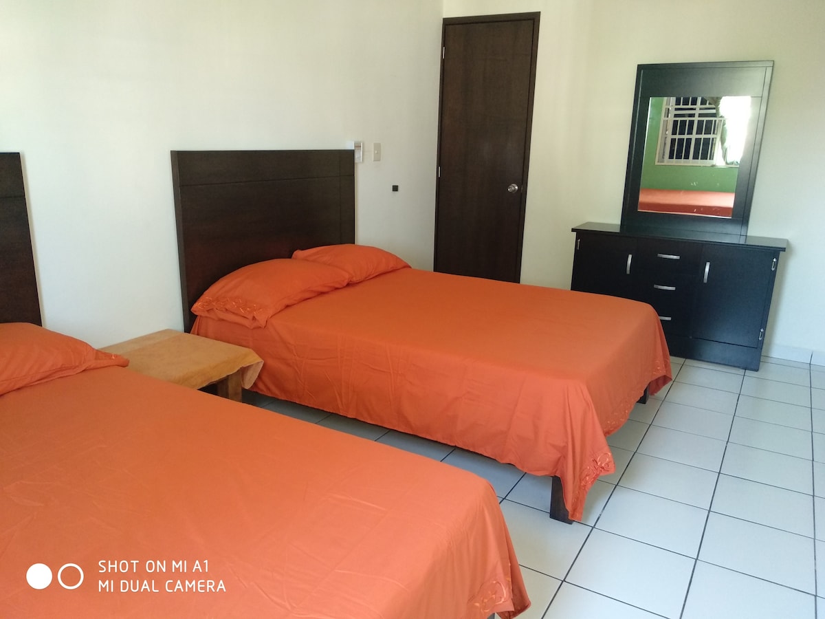 The image features a guest room with two orange beds positioned against the wall. A wooden nightstand is adjacent to one bed. A dark wood dresser with a mirror occupies the opposite wall, and large windows allow natural light to fill the space.