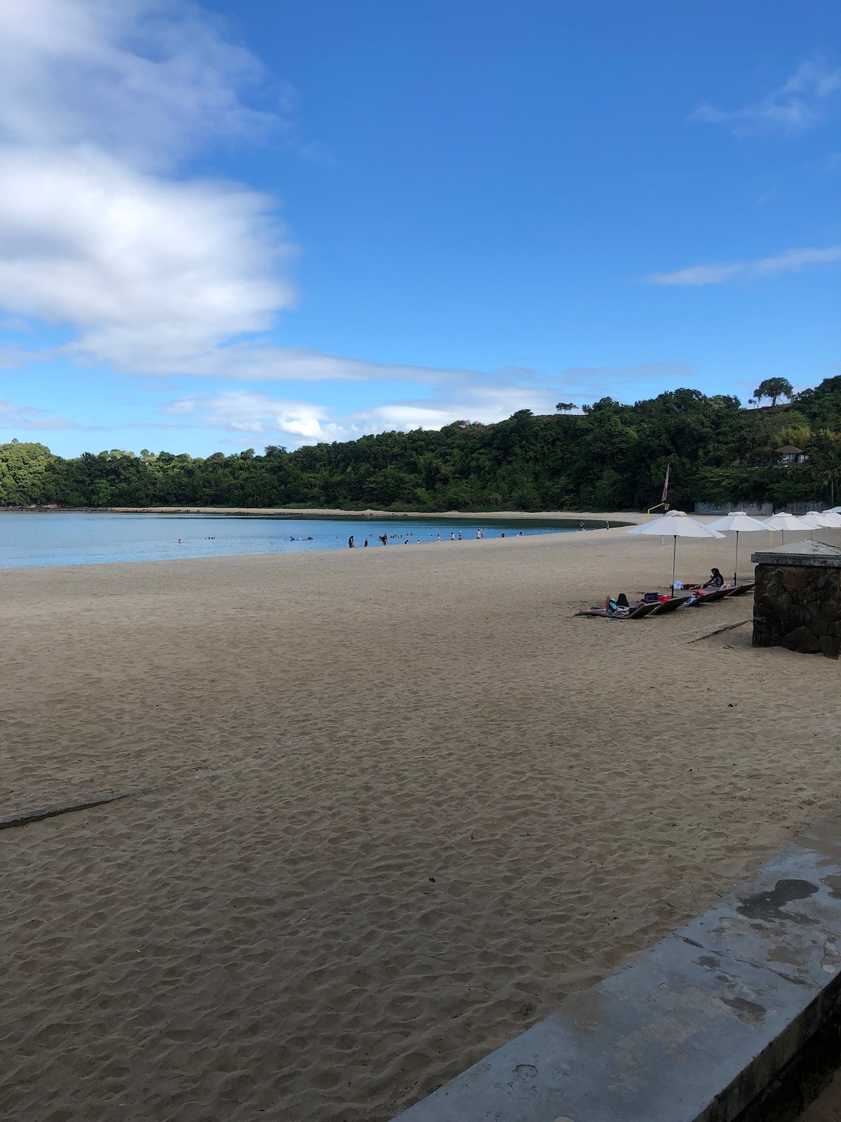 A sandy beach is framed by lush greenery and a calm body of water. Several beach loungers and umbrellas are positioned on the shore, while people can be seen enjoying the water in the distance under a clear blue sky with scattered clouds.