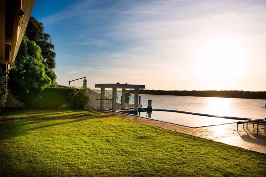 A sunlit outdoor area features a manicured lawn extending towards a tranquil river. A modern infinity pool reflects the sky, with a shaded seating area nearby. The scene captures the serenity of sunset over calm waters, providing a peaceful retreat environment.