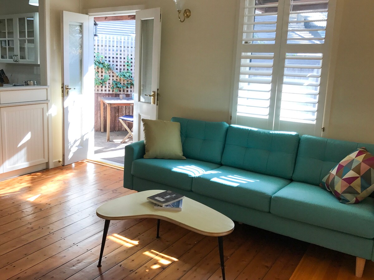 A light-filled living area features a turquoise sofa with a decorative pillow. A coffee table sits nearby, and large windows allow natural light to brighten the wood-paneled floor. A door leads to a private courtyard, enhancing the airy and open feel.