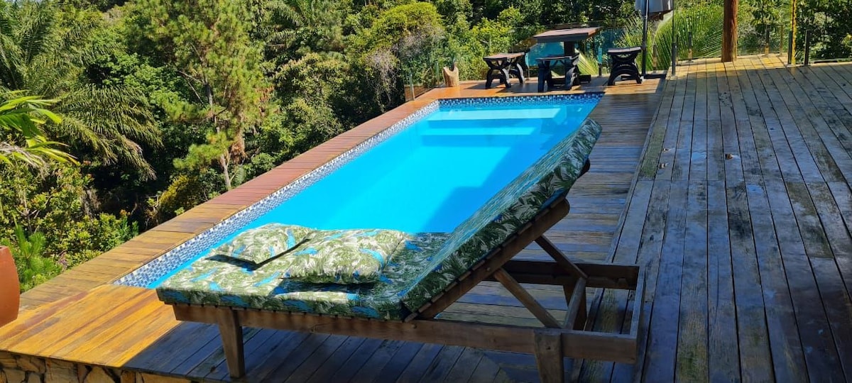 A refreshing swimming pool is framed by wooden decking, with lush green trees surrounding the area. A comfortable lounger rests beside the pool, adorned with a patterned cushion. Outdoor seating is visible in the background, inviting relaxation amidst the natural scenery.