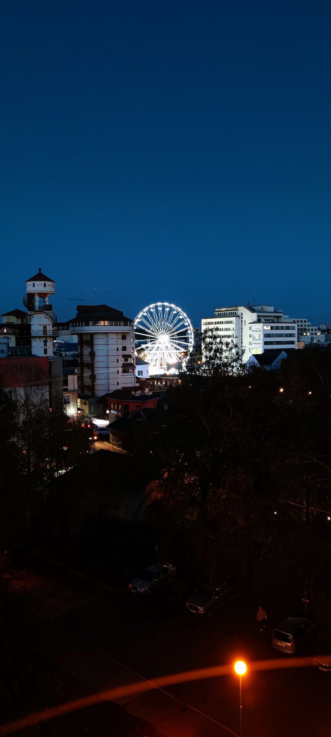 A nighttime cityscape is captured, showcasing illuminated buildings and a large Ferris wheel. The sky is dark blue, contrasting with the bright lights of the attractions. Nearby structures include a tower and various residential buildings, contributing to the urban atmosphere.