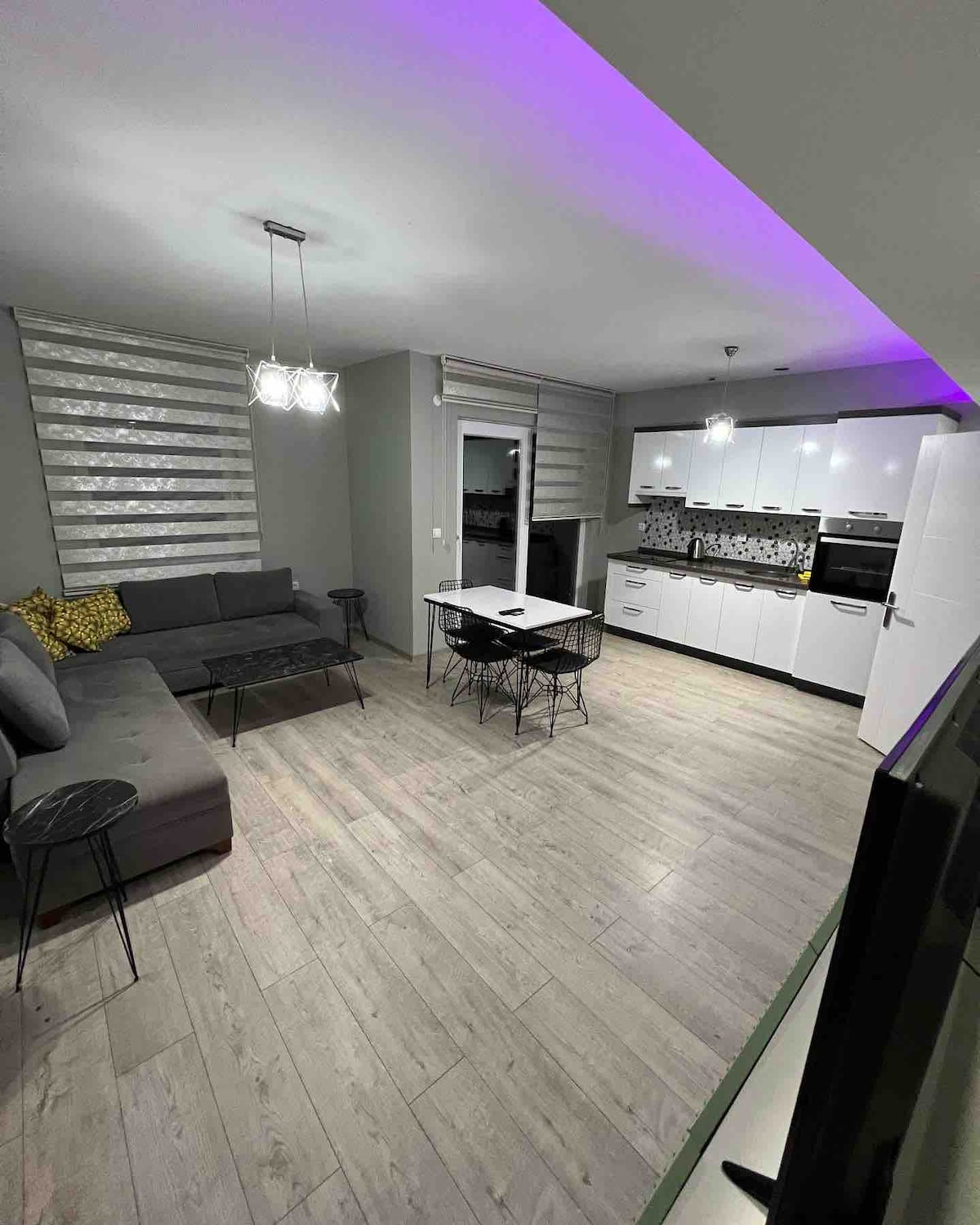 A modern living area features a sectional sofa and a glass-top coffee table. A dining table with black wire chairs is positioned nearby. The kitchen is equipped with white cabinetry and patterned backsplash, while soft lighting highlights the room's contemporary design.