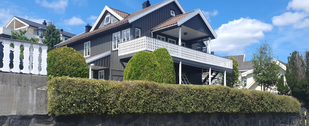 A charming two-story house with dark wood siding and a pitched roof is showcased. A spacious balcony with white railings spans the front, complemented by neatly trimmed hedges and a stone wall at the base. Lush greenery surrounds the property under a clear blue sky.
