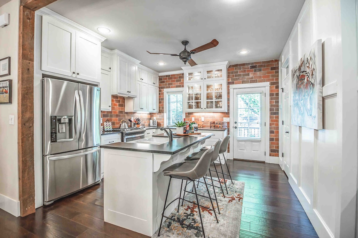 A modern kitchen features stainless steel appliances and a spacious island with seating for four. The design includes wood accents and exposed brick, enhancing the inviting environment. Natural light filters through windows, illuminating the room. A tasteful area rug adds warmth to the hardwood floor.