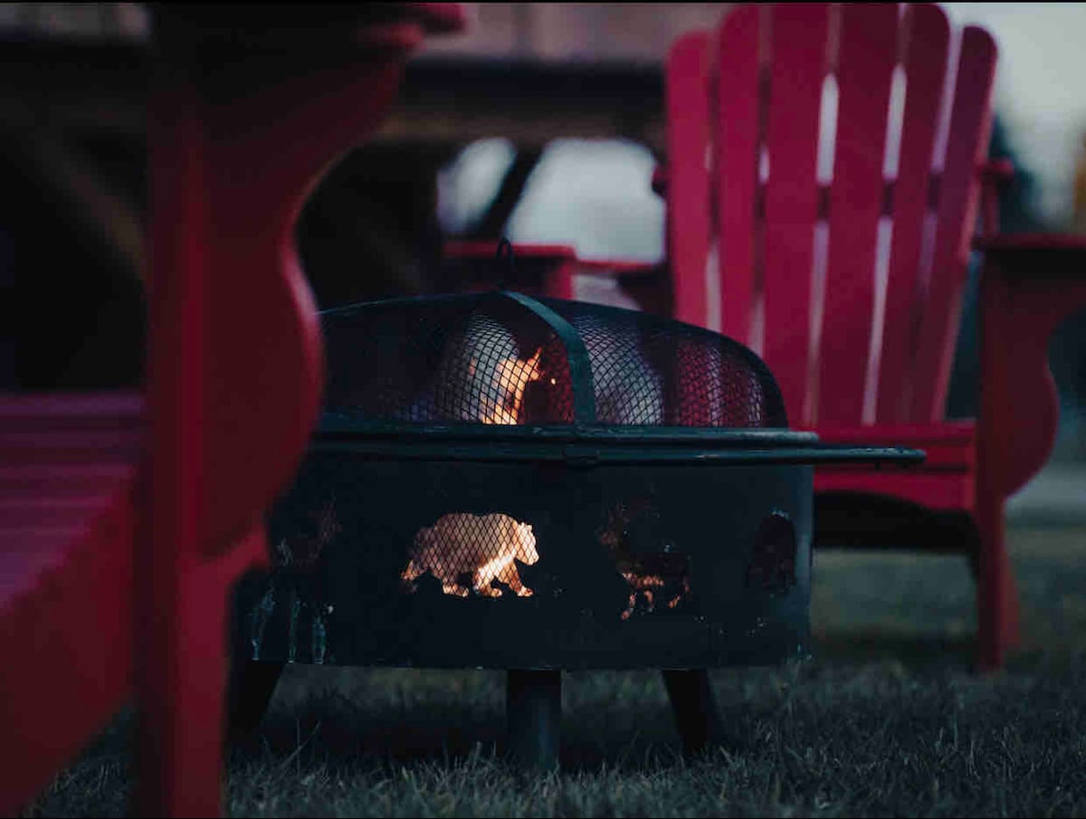 A fire pit is positioned in the outdoors, surrounded by two vibrant red Adirondack chairs. The flames flicker within a protective mesh cover, creating a cozy focal point in the tranquil setting.