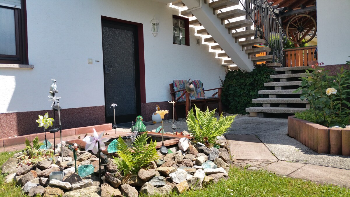 A charming outdoor area is featured with a decorative rock garden, showcasing a variety of glass sculptures among ferns and small plants. The entrance is framed by a staircase leading to an upper level, with comfortable seating visible beside the door.