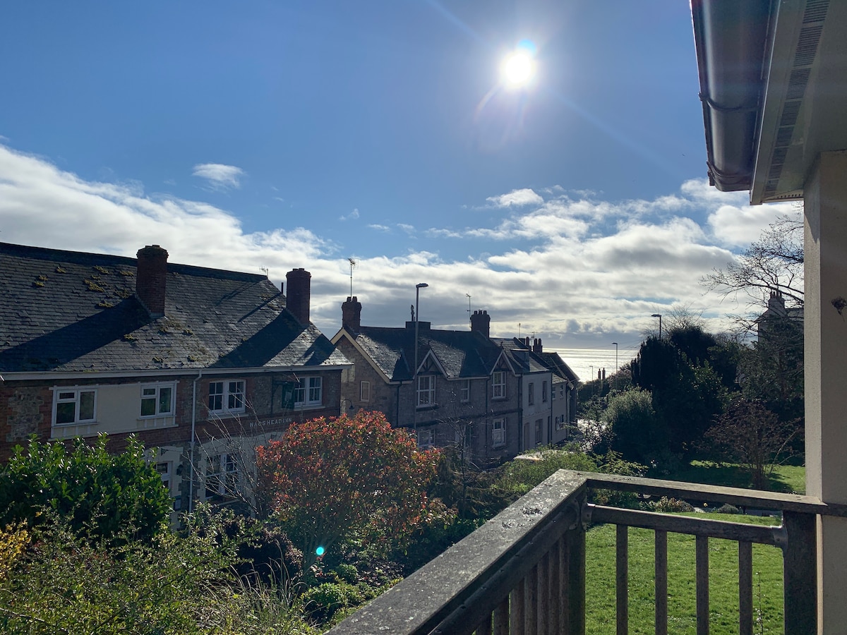 A view from a balcony features a clear blue sky with the sun shining above. Traditional homes are visible in the foreground, with garden greenery and a glimpse of the sea in the distance. Fluffy clouds create a serene backdrop.