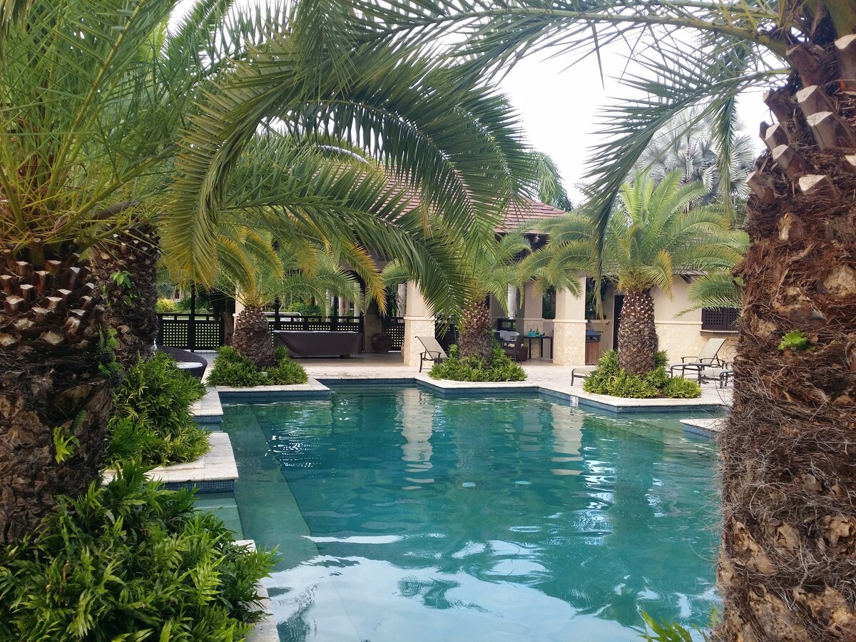 A tranquil pool area is surrounded by lush palm trees and tropical greenery. The clear water reflects the surrounding foliage, while a covered seating area is visible in the background. The setting offers a peaceful oasis for relaxation.