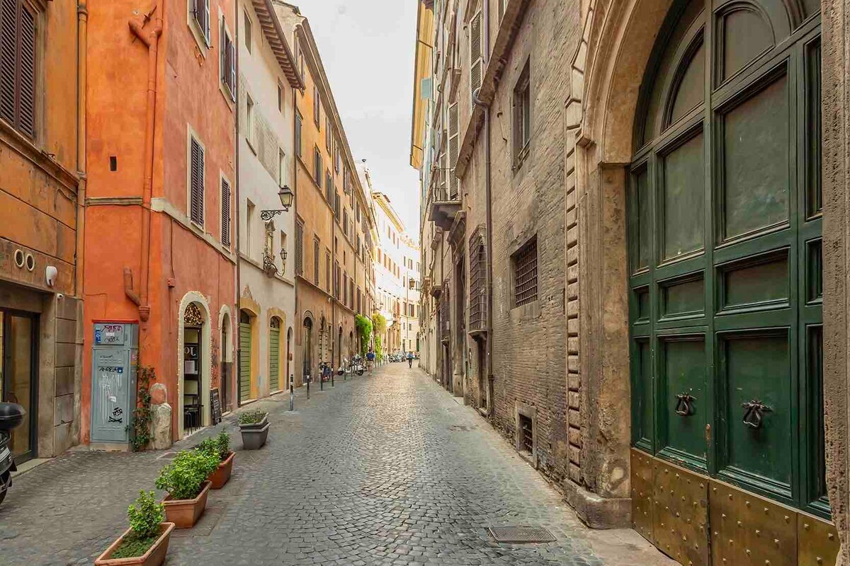 A narrow cobblestone street is flanked by charming, colorful buildings with various window styles. The architecture displays a blend of textures and colors, including warm orange and muted brown tones. Planters with greenery are positioned along the sidewalk, creating a welcoming ambiance.