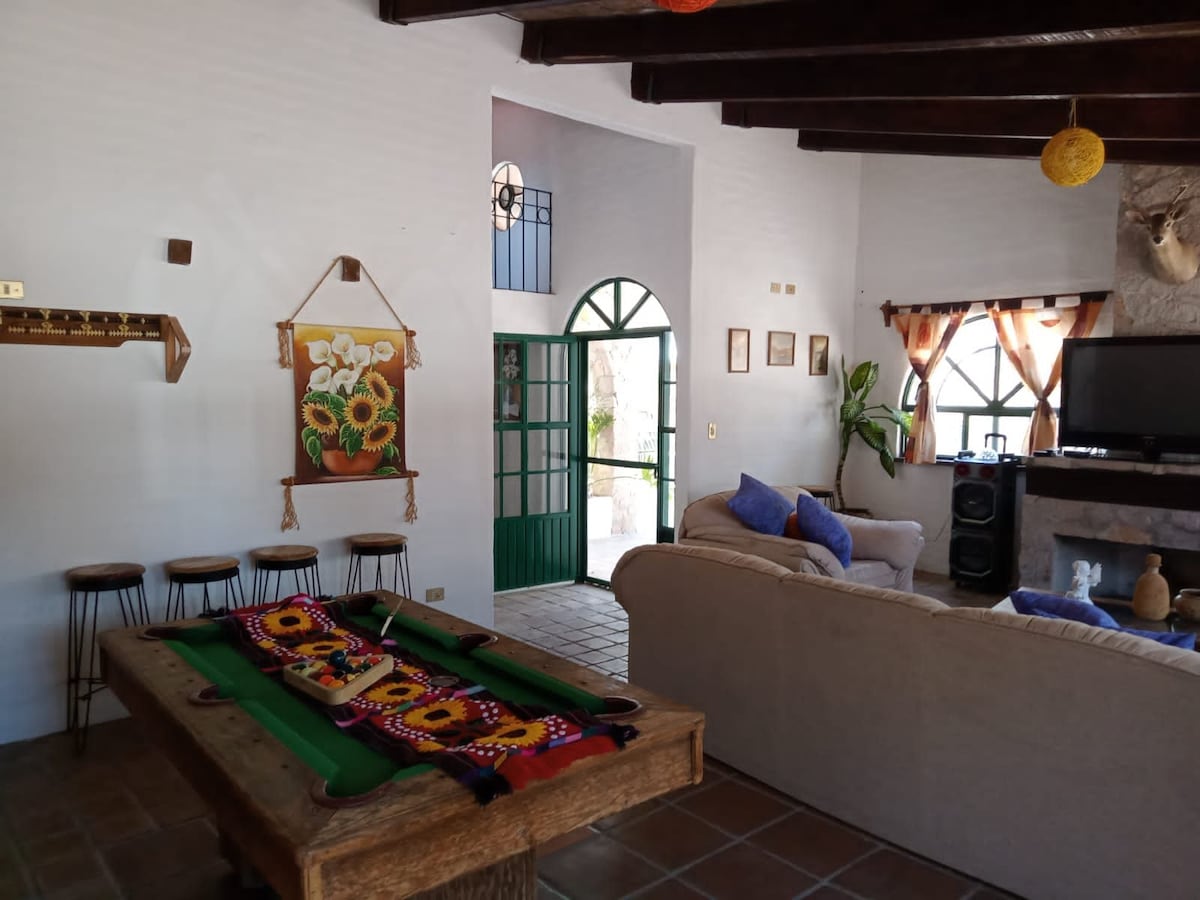 An open living area features a rustic wooden pool table adorned with colorful cloth. A beige sofa is positioned across from a television, complemented by a traditional fireplace. Light streams through the arch-shaped green doorway, while art and decor add charm to the inviting space.