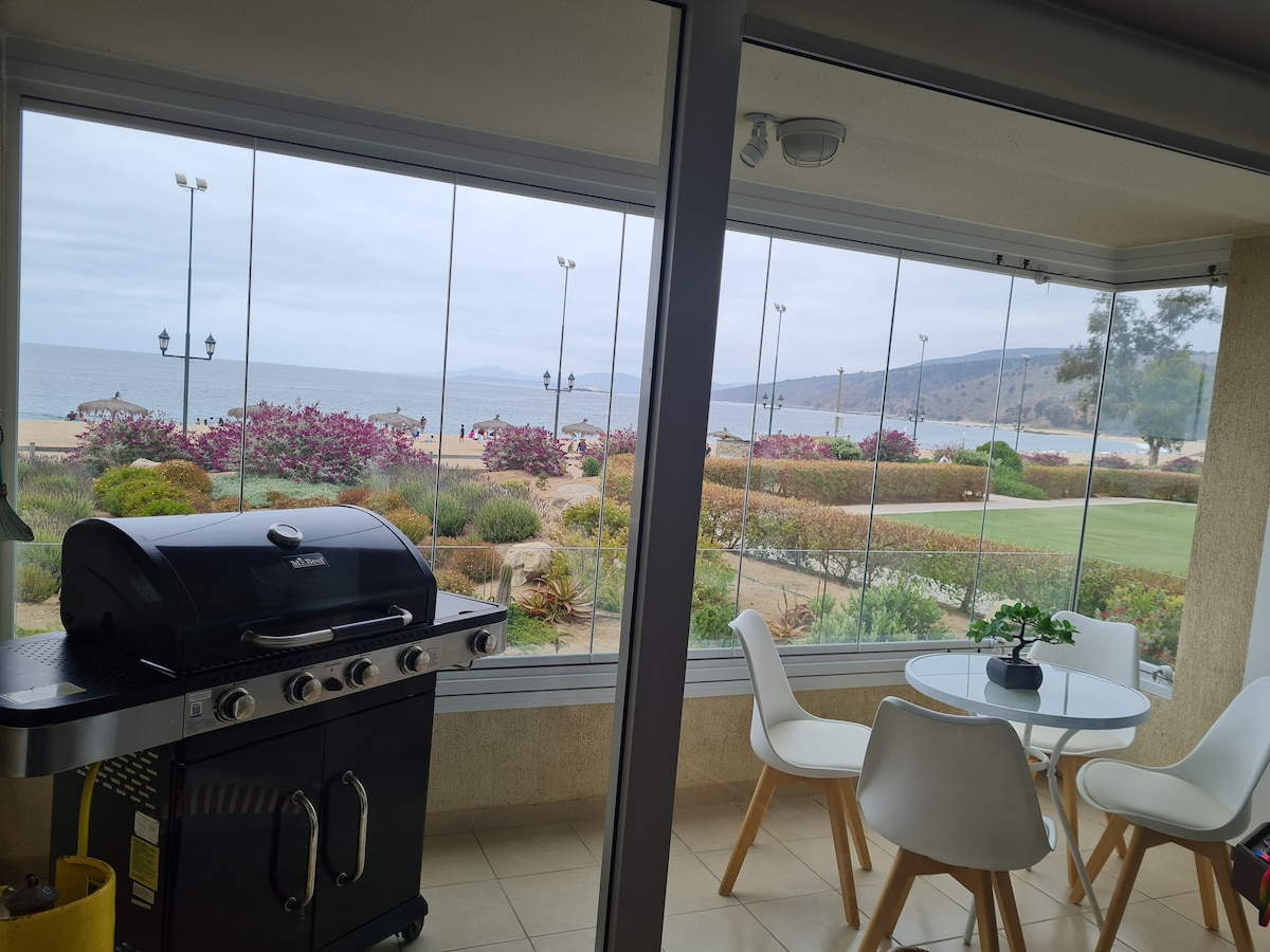 A grill is positioned next to a round dining table with four chairs, overlooking a sandy beach and ocean view through large glass windows. Colorful landscaping surrounds the area, and a path is visible in the distance where people can be seen enjoying the outdoor space.