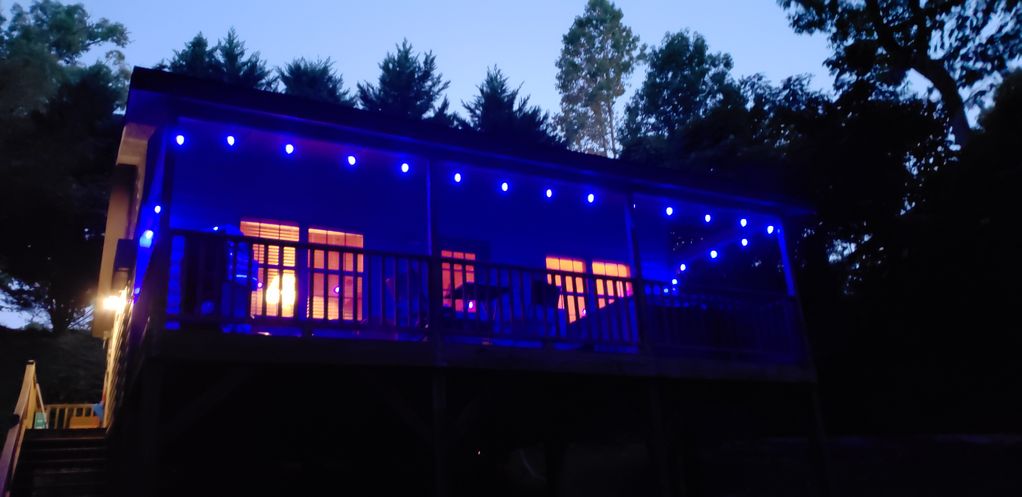 A rustic cabin is seen at dusk, featuring soft blue lighting along the deck and near the entrance. The warm glow from the windows creates a welcoming appearance, surrounded by trees that provide a sense of privacy. The structure is elevated on stilts, enhancing the outdoor presence.