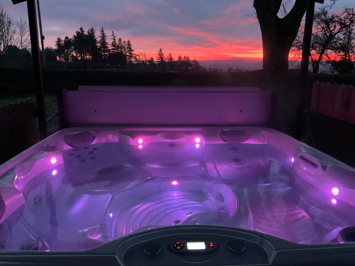A private hot tub, illuminated with soft purple lighting, is positioned next to the cottage. The scenic backdrop displays a vibrant sunset with hues of orange and pink, framed by silhouette trees on the horizon.