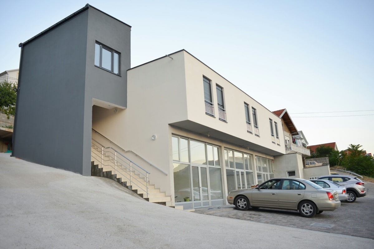 The modern exterior of the motel displays a two-tone color scheme, featuring a sleek grey wall and large glass windows that allow natural light into the building. A gently sloping driveway leads to a parking area, accommodating several vehicles.