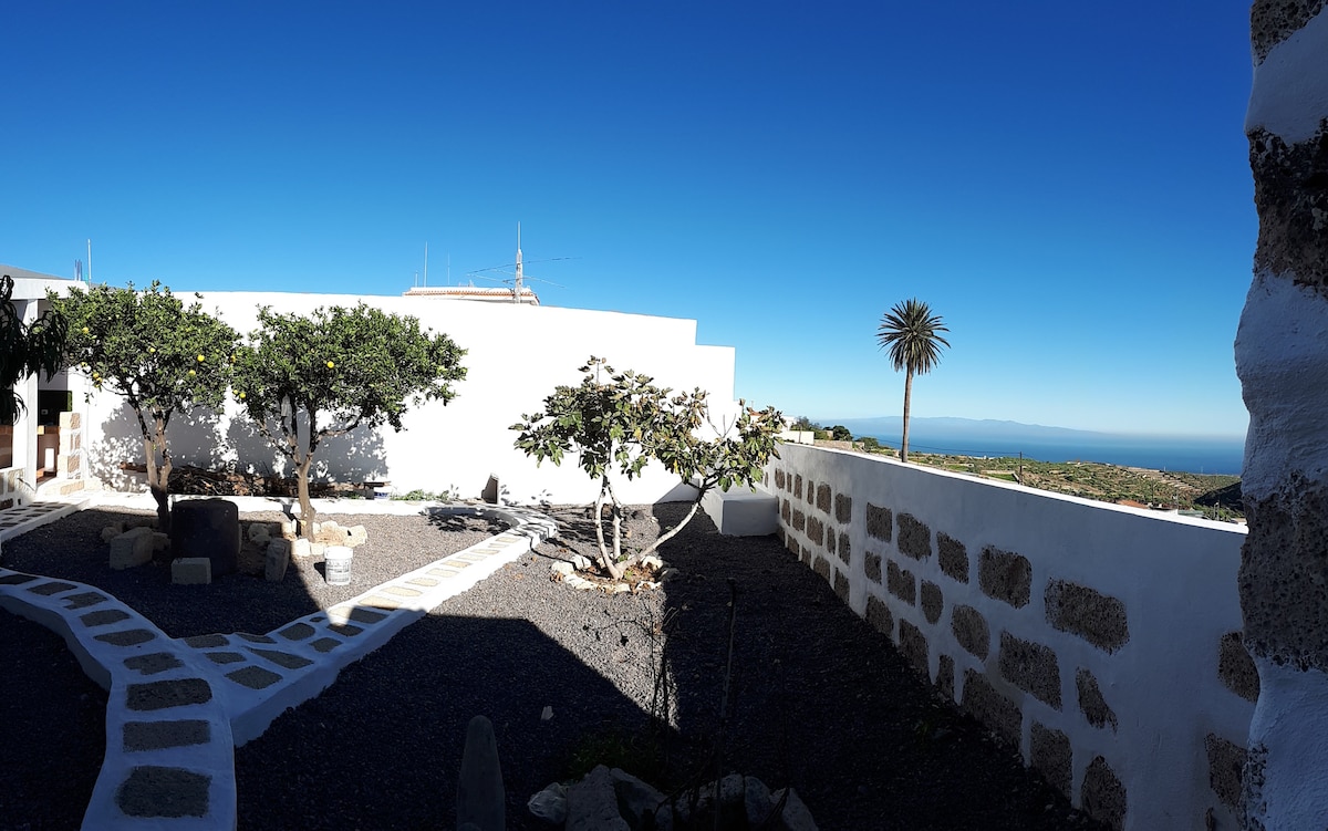 A spacious garden area features a stone pathway surrounded by various trees, including orange trees and palm. The clear blue sky complements the white walls of the building in the background, while the sea is visible on the horizon, enhancing the outdoor setting.