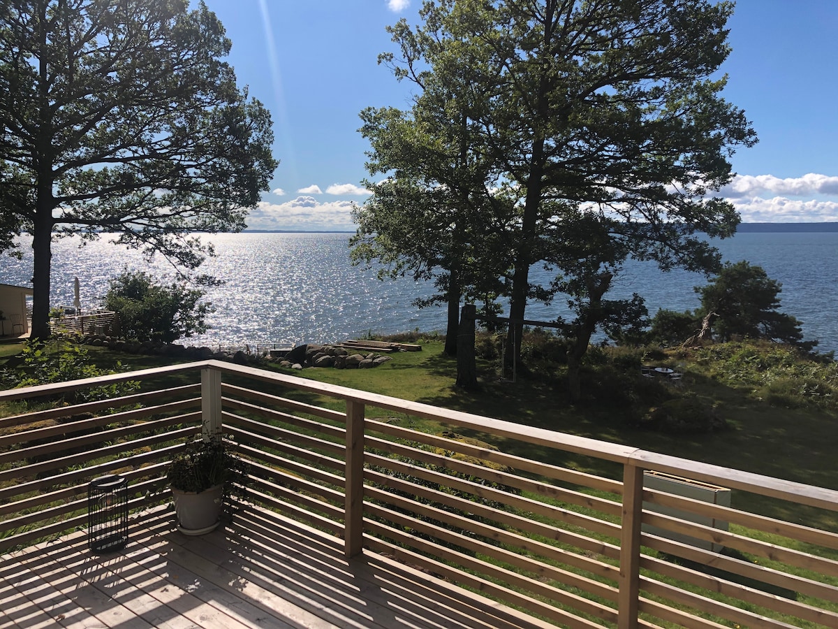 A wooden deck overlooks a shimmering body of water, surrounded by lush greenery. Tall trees provide shade, and distant views of a natural shoreline are visible. The scene is bathed in natural light, creating a peaceful and serene environment.