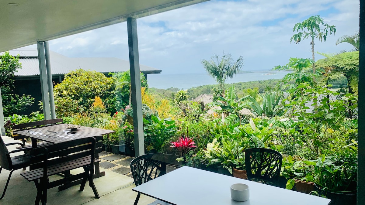 A shared balcony showcases lush tropical greenery alongside seating arrangements. Ocean views are visible in the distance, framed by the vibrant plants. The outdoor area offers a relaxing space for guests to enjoy the scenery and fresh air.