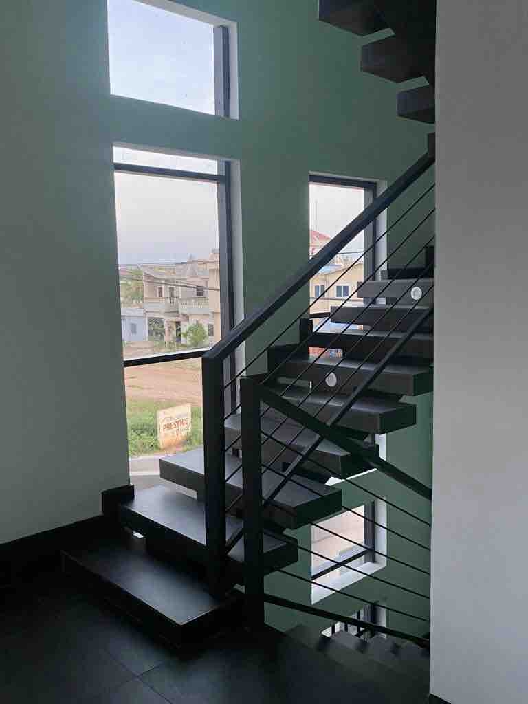 A modern staircase is viewed from a lower level, featuring sleek metal railings and wooden steps. Large windows allow natural light to illuminate the space, framing views of the surrounding area. The walls are painted in a calming light color, contributing to the overall serenity.