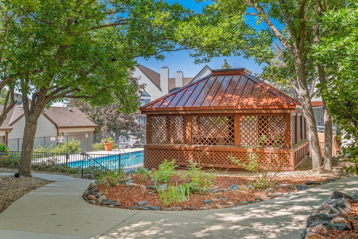 A shaded gazebo with a wooden structure is surrounded by lush greenery and decorative landscaping. The pathway leads to the inviting pool area visible in the background, where sun loungers are positioned for relaxation.