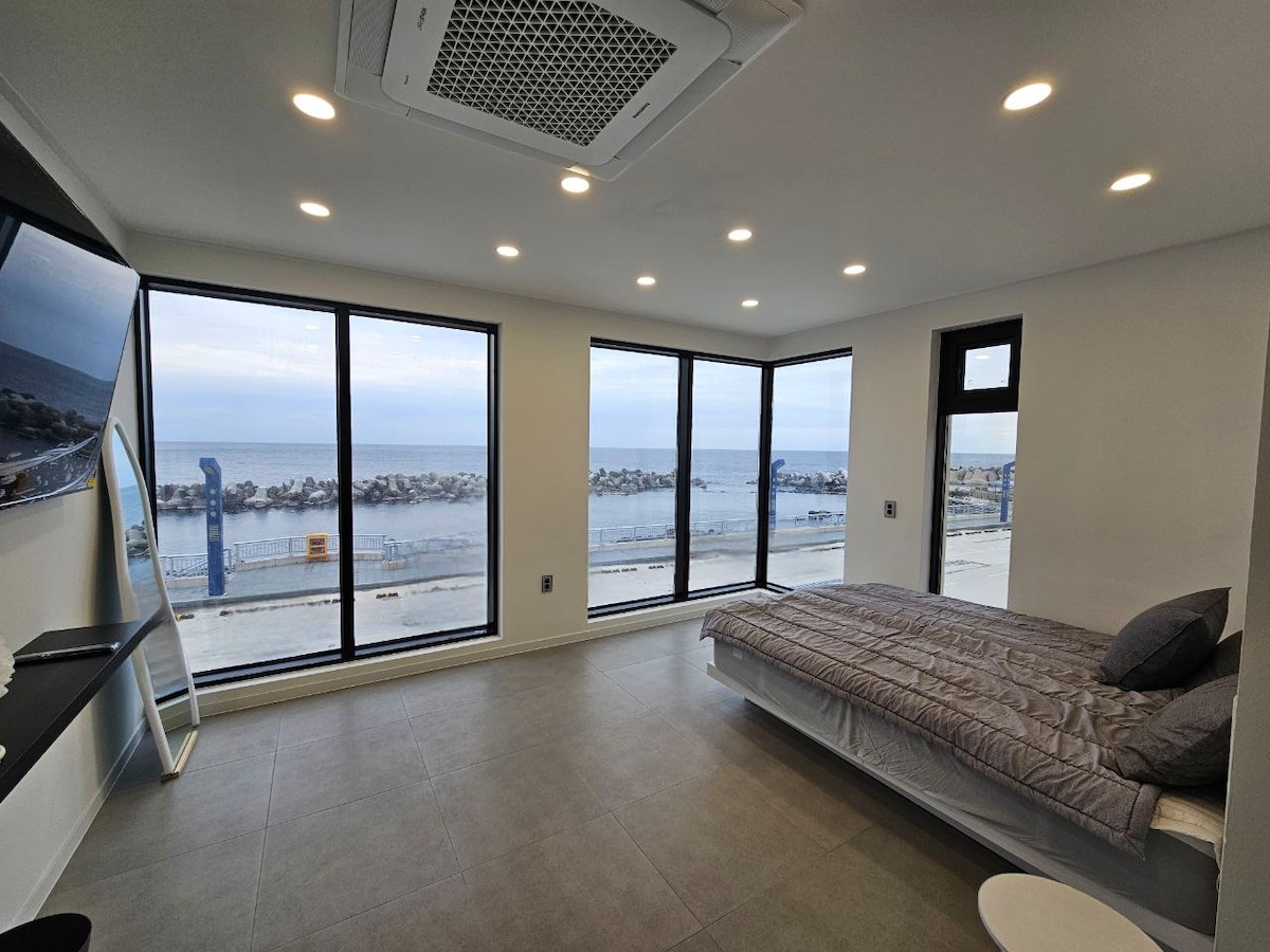 A bright and airy room features floor-to-ceiling windows offering a stunning view of the ocean and rocky shoreline. A spacious bed is positioned against one wall, complemented by a modern television unit. Soft lighting creates a serene environment throughout the space.