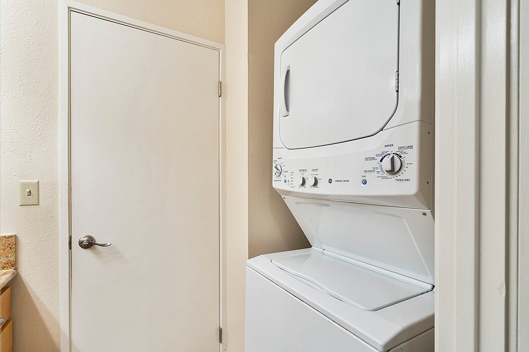 A stacked washer and dryer unit is positioned against a beige wall within a small laundry space. A white door is visible nearby, providing entry to another room. Counter space with granite countertops is partially seen to the left.