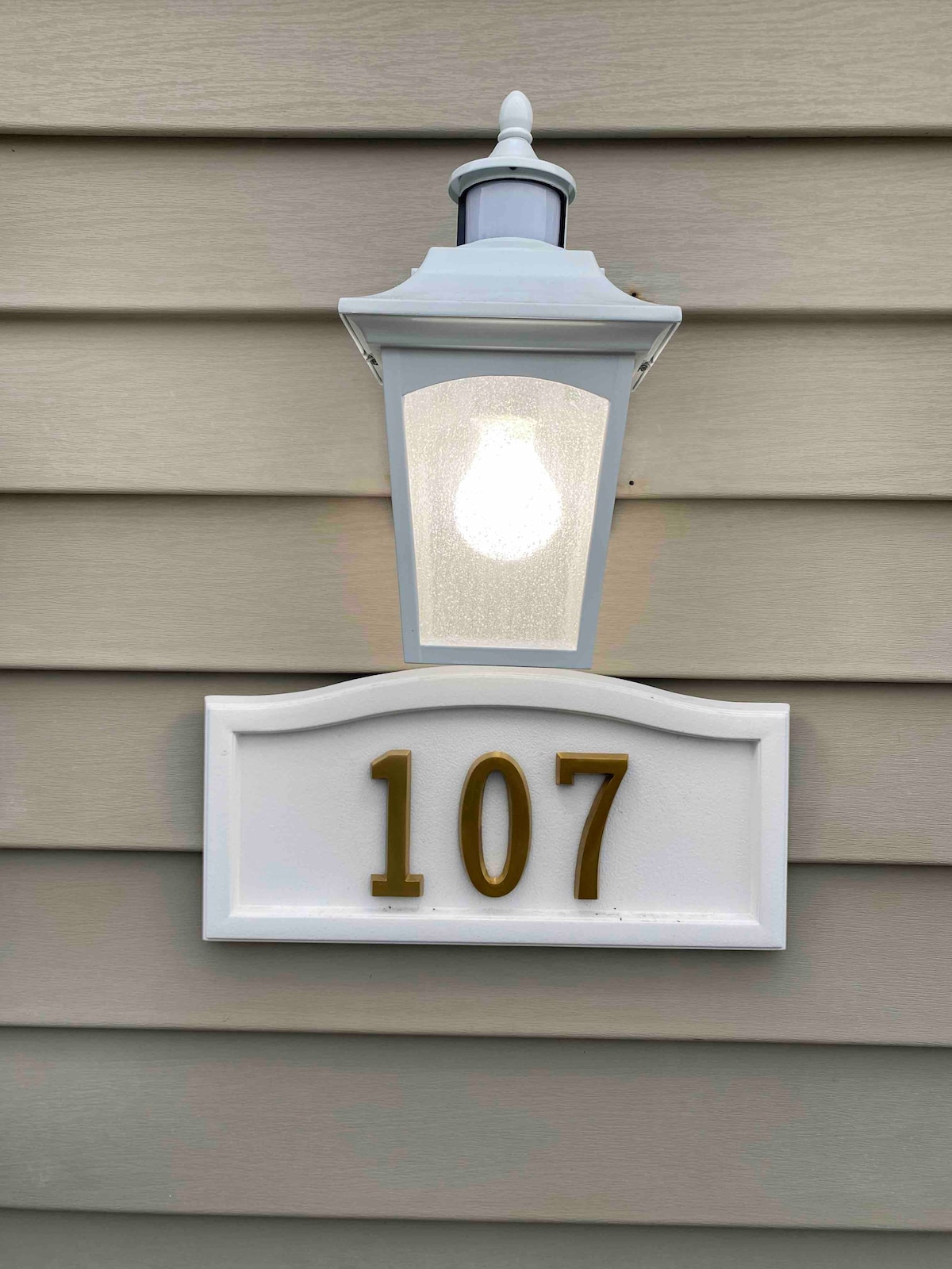 The image displays a white lantern-style light fixture mounted on a textured exterior wall. Below the light, a white plaque is visible, featuring the number '107' in gold. The soft illumination from the light adds clarity to the house number.