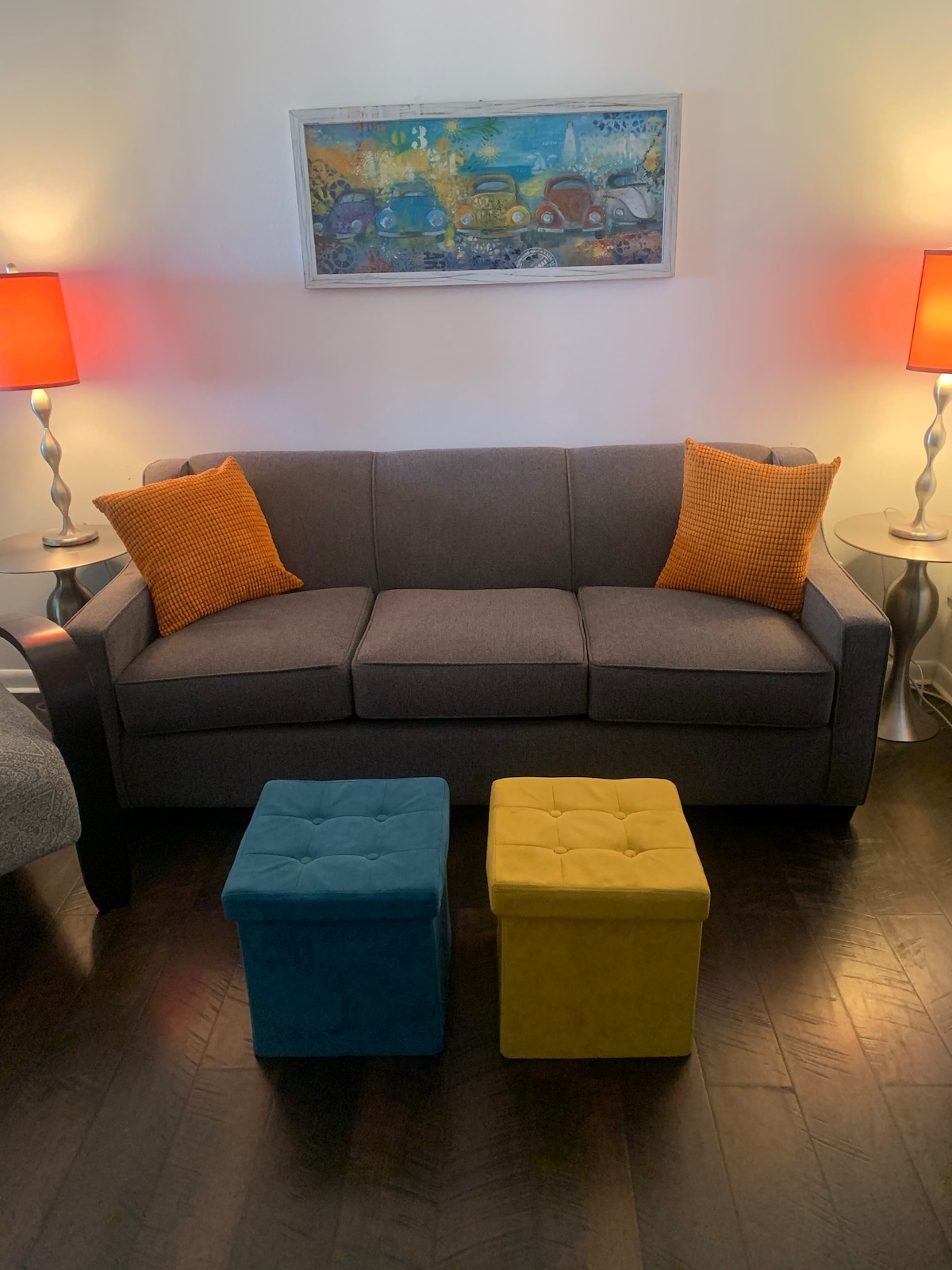 A cozy seating area features a gray sofa adorned with two orange accent pillows. Flanking the sofa are two colorful ottomans, one in blue and the other in yellow. Two stylish lamps with red shades provide soft lighting, while a framed artwork hangs above the sofa.