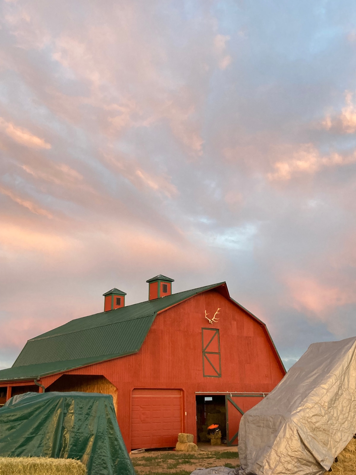 A red barn stands prominently against a sky painted with soft pastel hues at sunset. Two cupolas adorn the roof, adding architectural interest. Bales of hay are covered nearby, contributing to the rustic farm atmosphere.