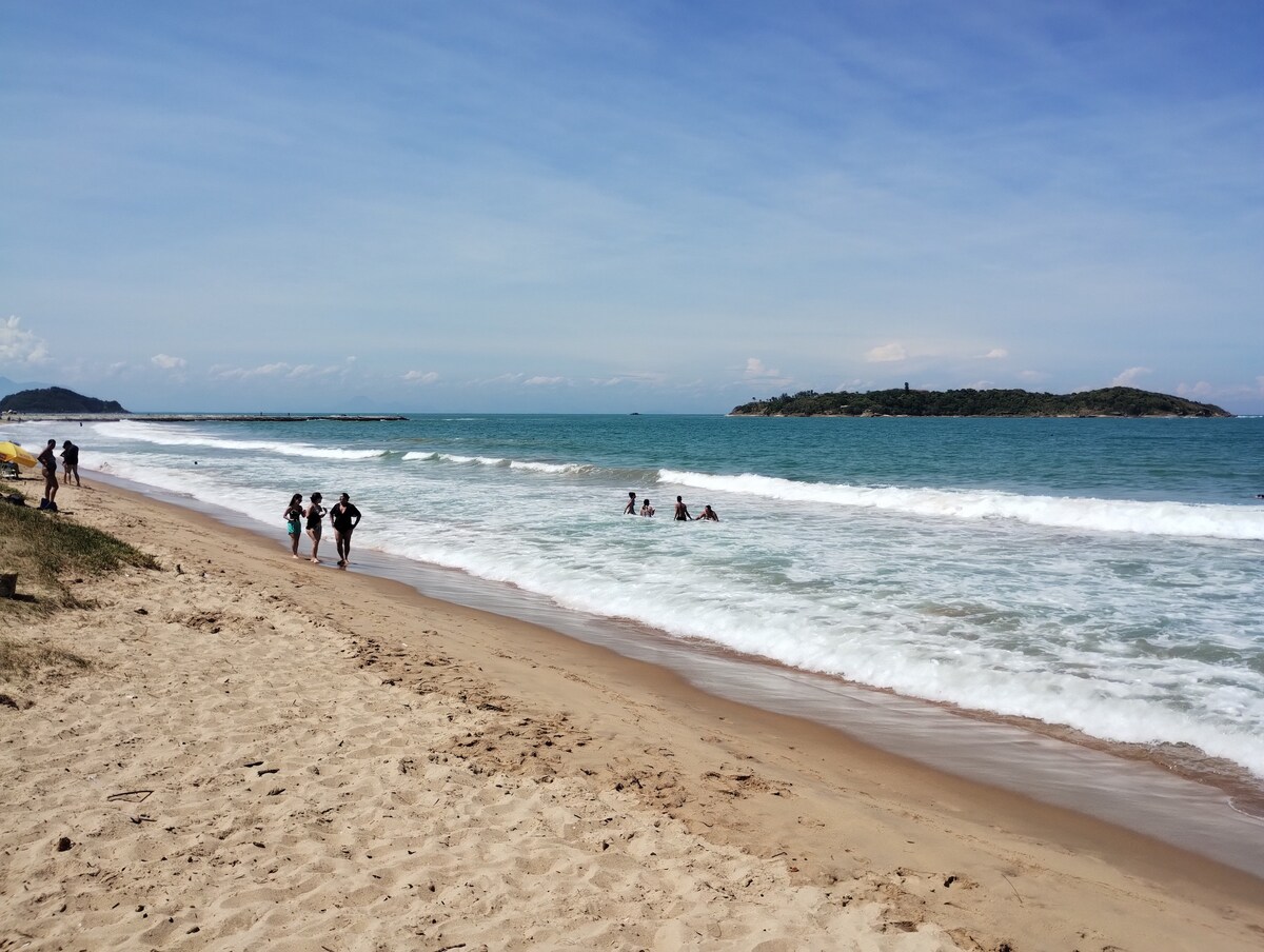A sunny beach scene is depicted, featuring gentle waves lapping the shore. Several individuals can be seen walking along the sandy beach, while others enjoy the water. A distant island is visible on the horizon, adding to the coastal landscape.