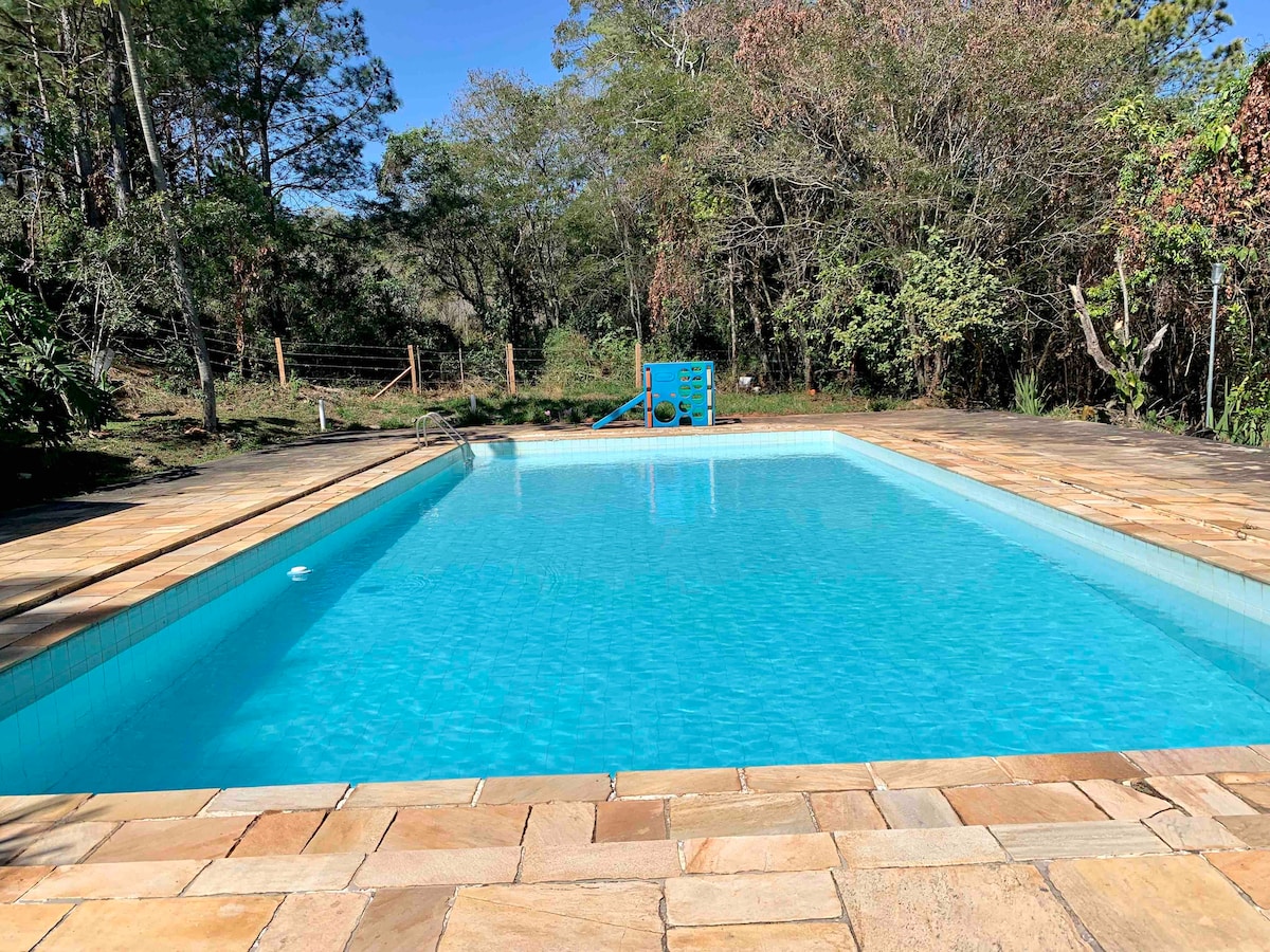 A serene swimming pool is surrounded by natural greenery, with light blue water reflecting the clear sky. The pool features smooth stone decking, and a children's play area can be seen in the background, blending harmoniously with the tranquil environment.