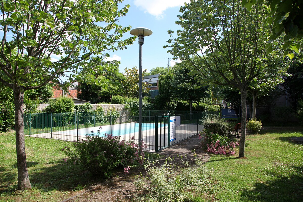 A tranquil garden area features lush green trees and blooming flowers surrounding a fenced swimming pool. The pool is situated at the center, with a light pole nearby, creating a serene outdoor space ideal for relaxation.